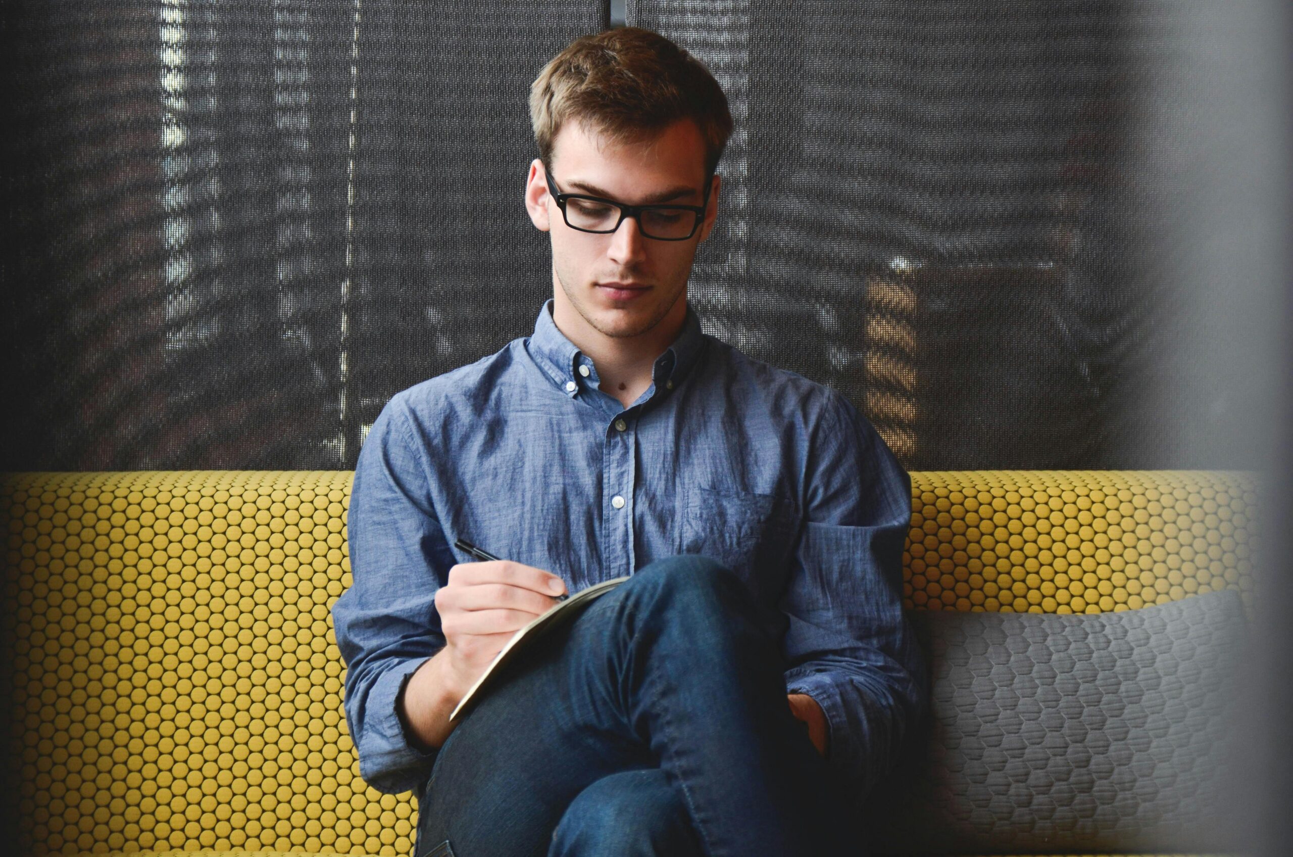 O NAS A young man in glasses writes in a notebook while sitting on a stylish couch indoors.